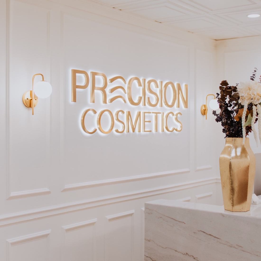 Precision Cosmetics reception — gold signage on a panelled cream wall with brass sconces and a marble desk